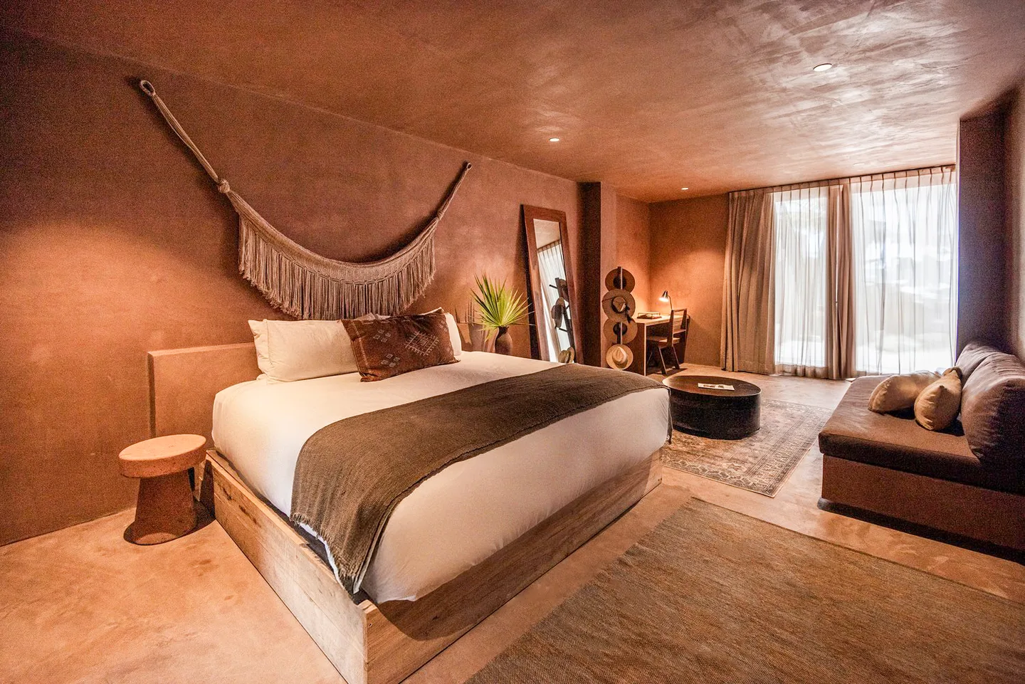 King bed beneath a woven hammock wall hanging, with the lounge area and window beyond