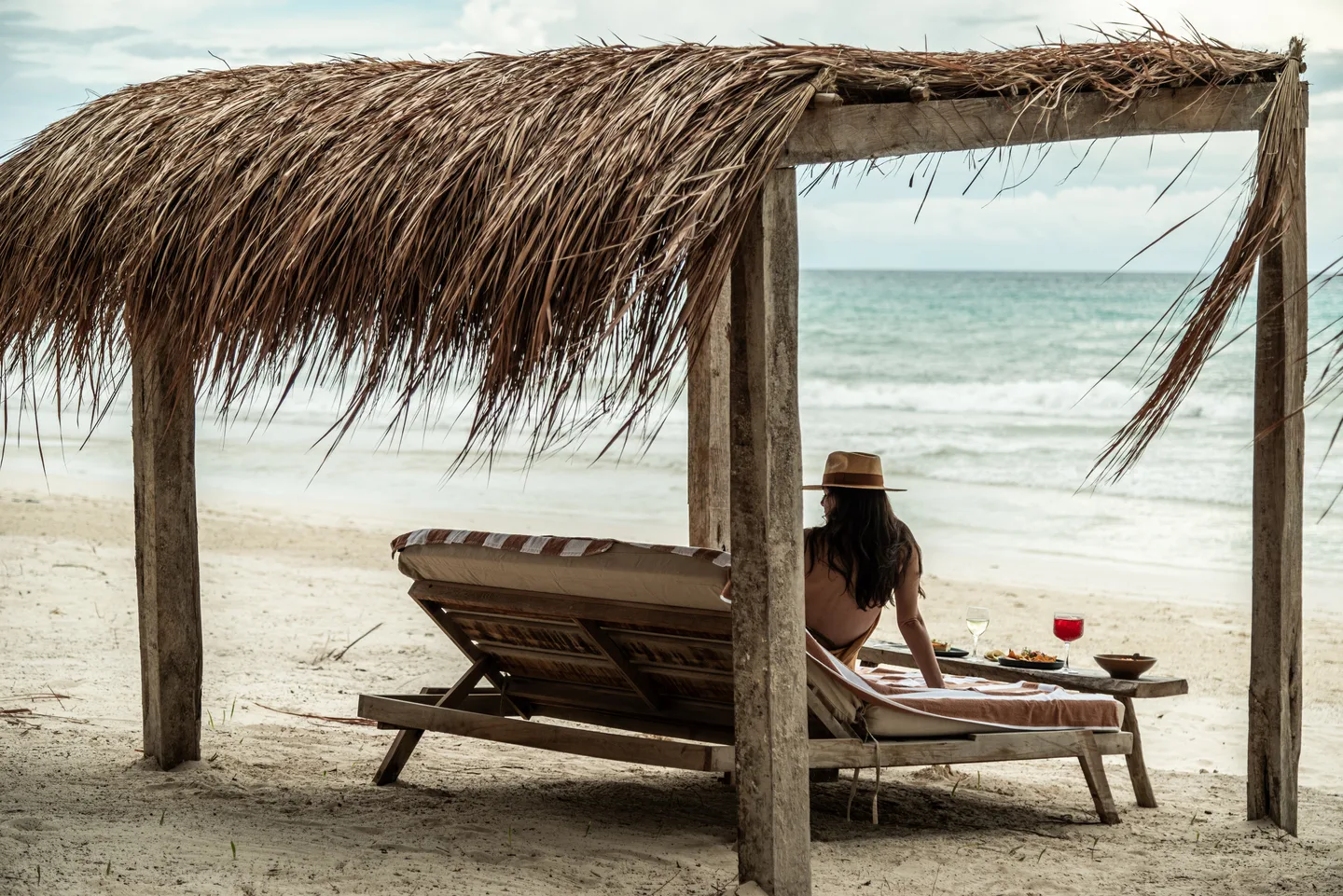 Guest on a lounger under a thatched palapa looking out to sea, lunch and wine beside her