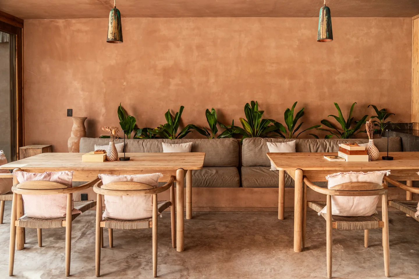 Restaurant dining room with light wood tables, cushioned banquette and a planter of tropical leaves