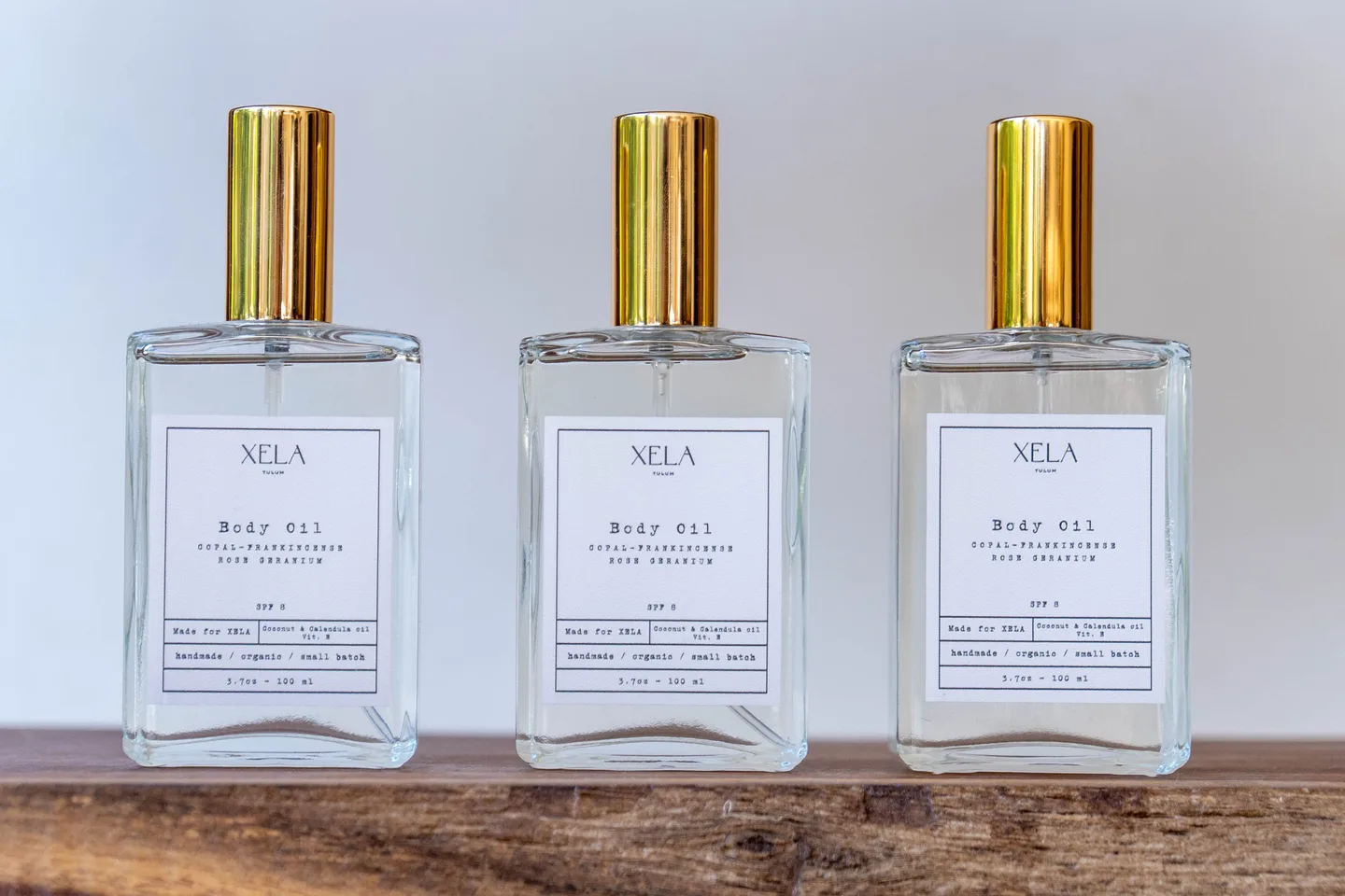 Three bottles of XELA body oil with gold caps on a wooden shelf