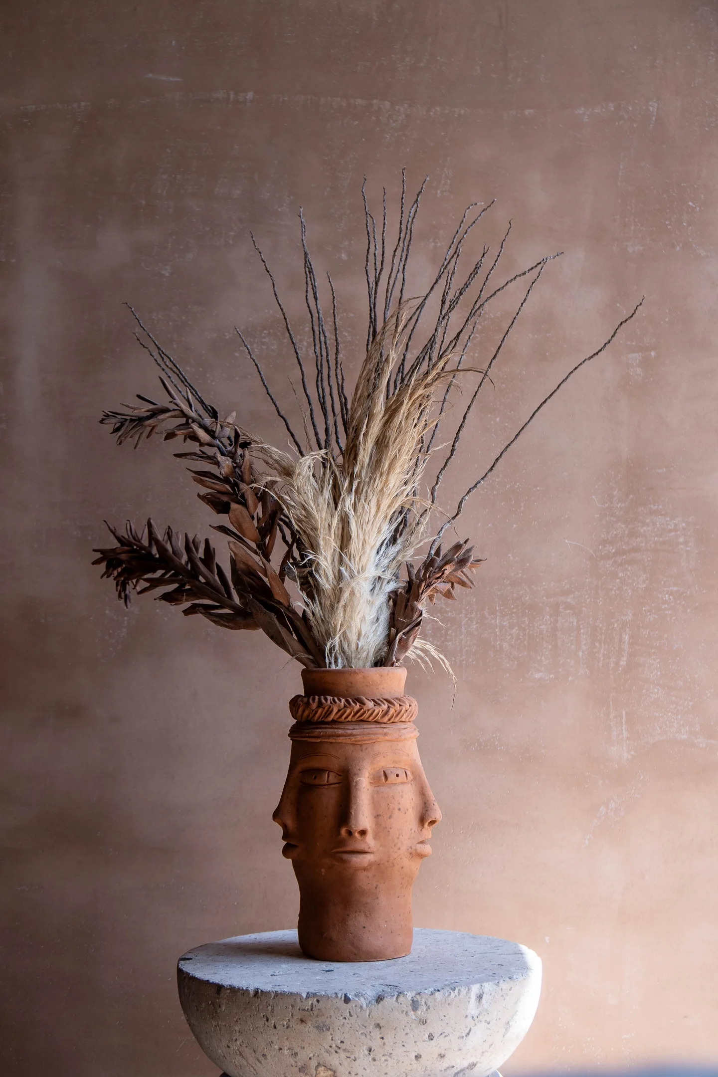 Clay face vase holding dried grasses on a stone pedestal