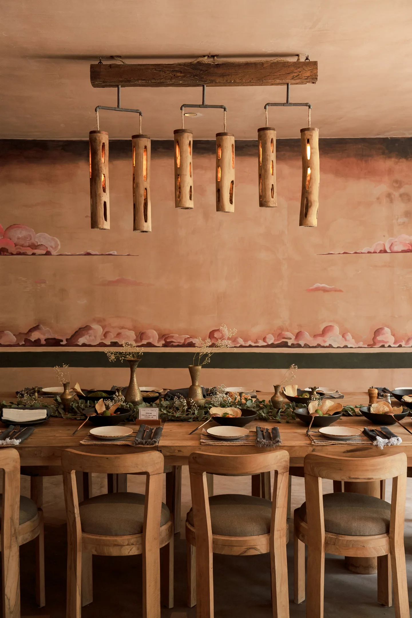Private dining table dressed with greenery beneath wooden pendant lights and a painted mural